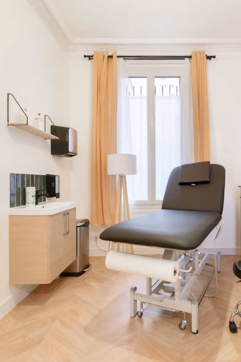 Salle de consultation médicale avec une table d'examen noire, un lavabo avec distributeur de savon, une poubelle et une grande fenêtre avec rideaux beiges.